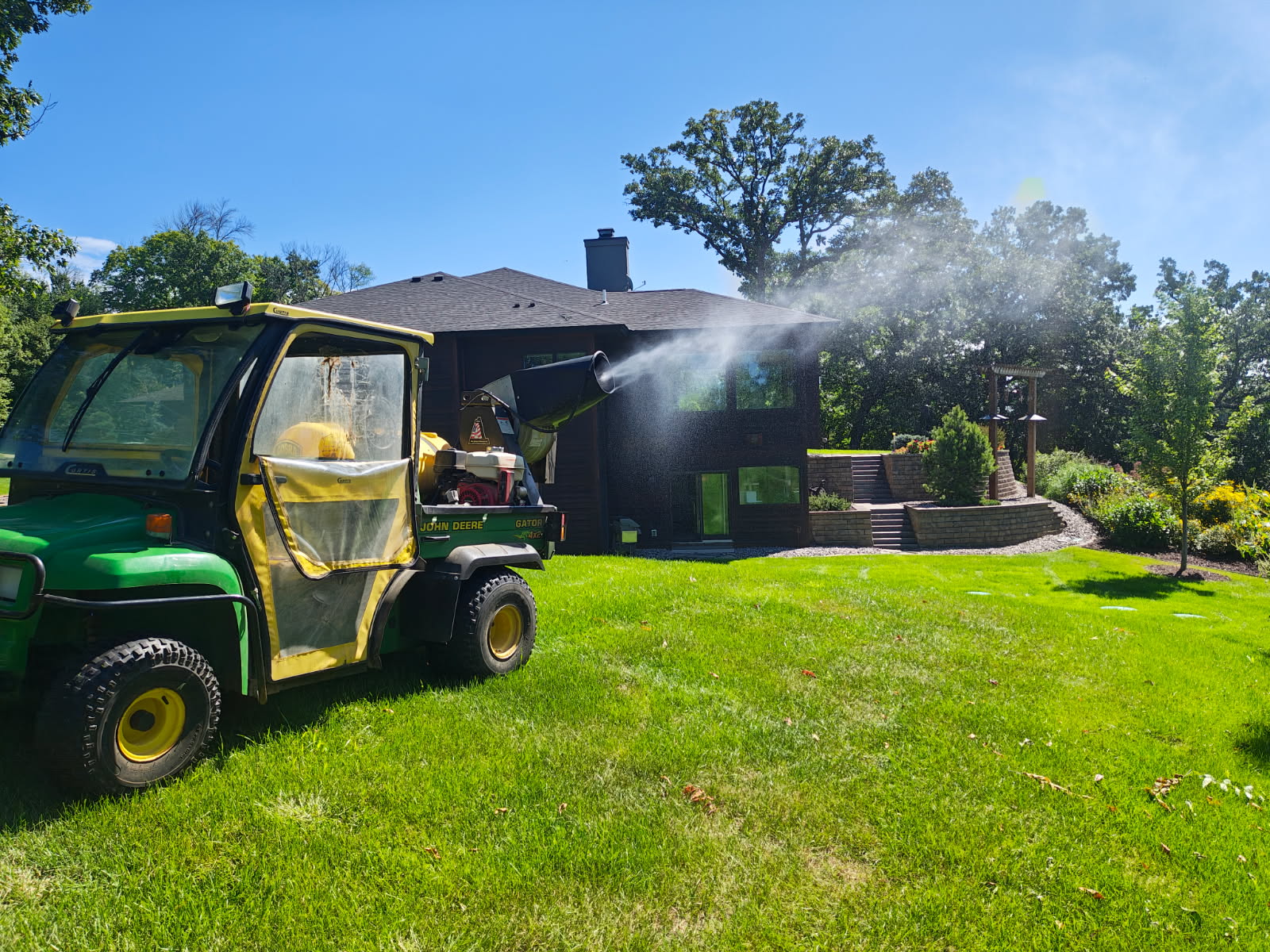 Professional mosquito control treatment being applied on a residential property in Central Minnesota, with mosquito fogging equipment treating a lawn, landscape beds, and shaded outdoor areas.